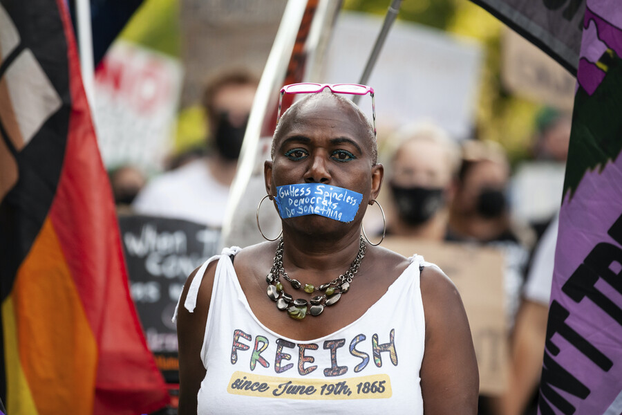 A person standing in a crowd wearing a piece of tape over their mouth that reads, "Gutless, spineless Democrats, Do Something," wearing a shirt that reads, "FREEISH since June 19th, 1865"