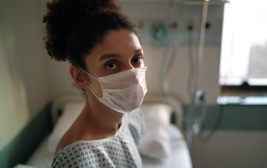 A transgender patient wearing a mask and hospital gown on a hospital bed.