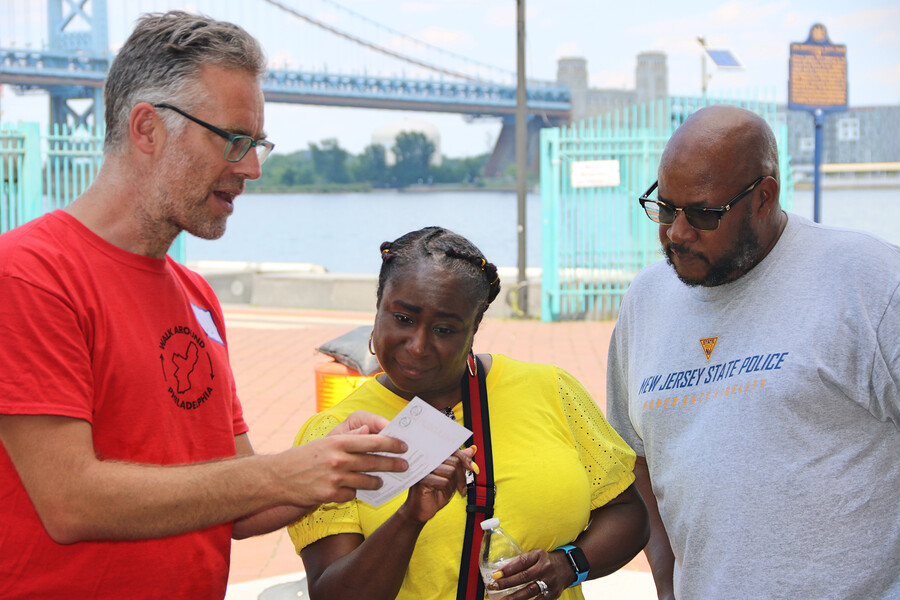 Three people standing on an outdoor walkway with the Ben Franklin Bridge in the background.