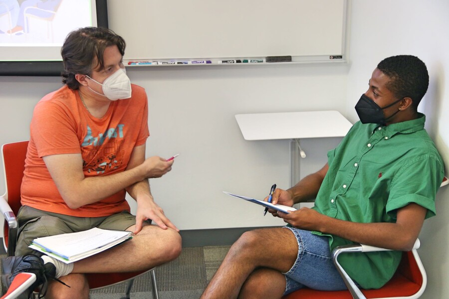 Two men in KN-95 masks sit in attentive discussion, one wears an orange shirt and the other wears green.