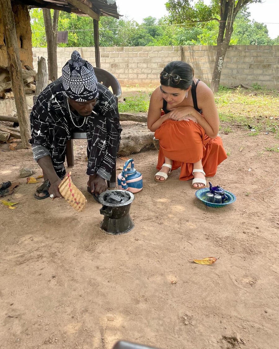 A man in a black, patterned outfit sits and stokes coals next to a woman squatting in an orange skirt.