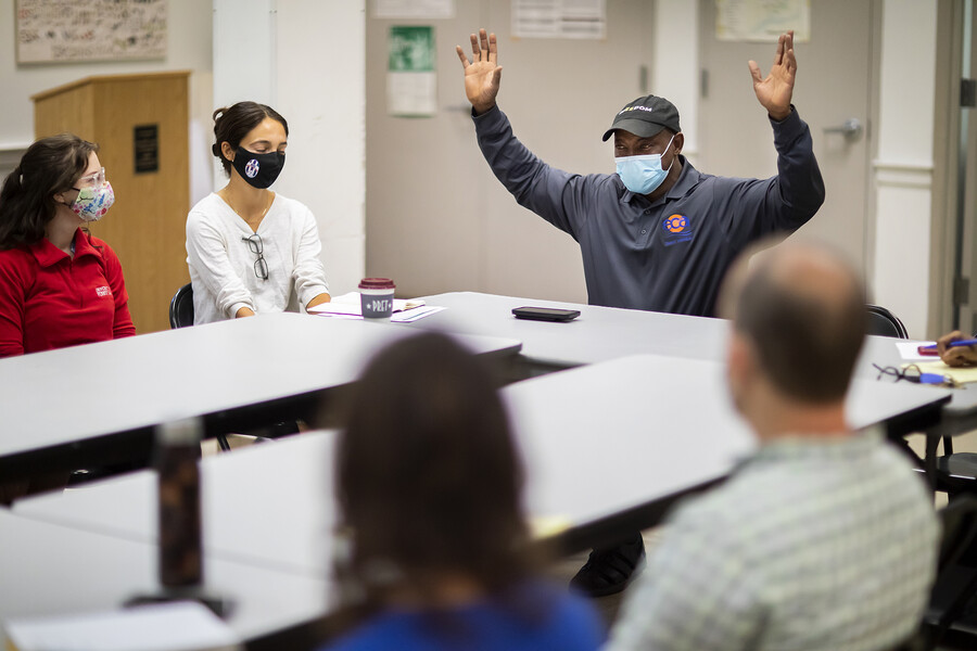 Five people seated at a table wearing masks, Ocek Eke at head of table raising their hands.