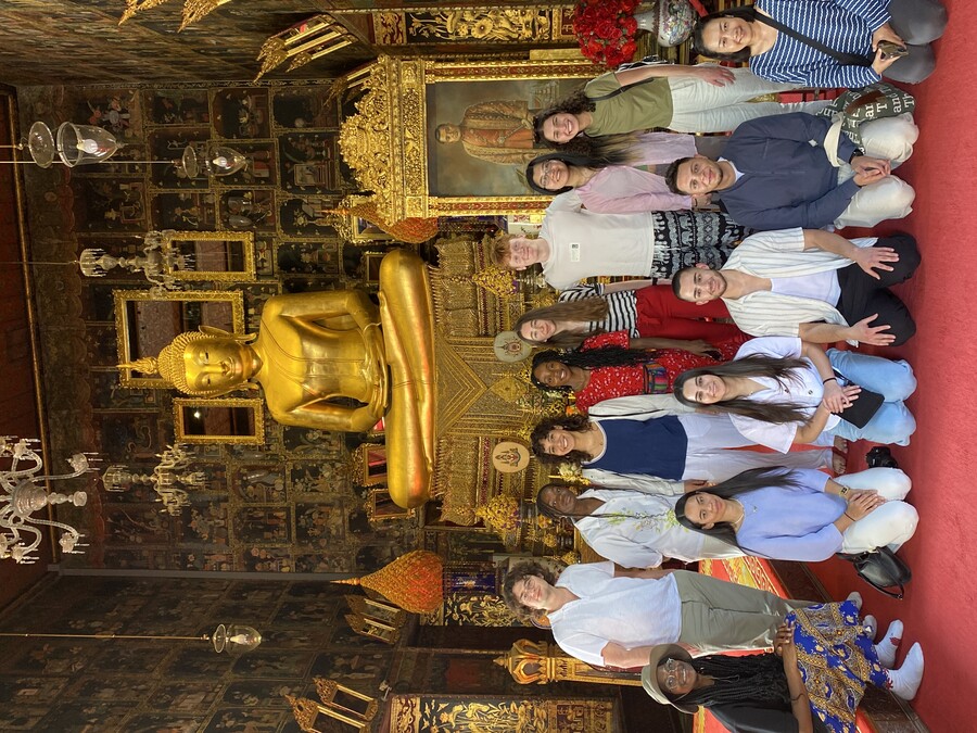 Students pose in the interior of a Buddhist temple, with a large Buddha and hanging light fixtures 