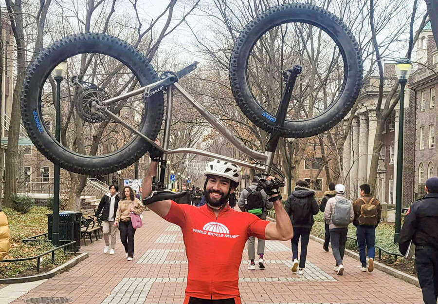 Ryan Torres holds his mountain bike aloft on Locust Walk in cycling gear.