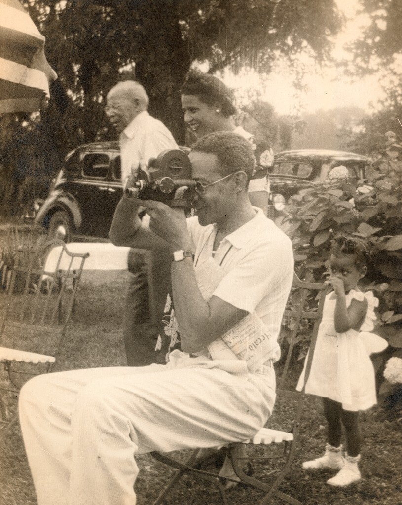 Raymond Alexander holding a movie camera while sitting in a chair outside with cars and flowering bushes behind him. 