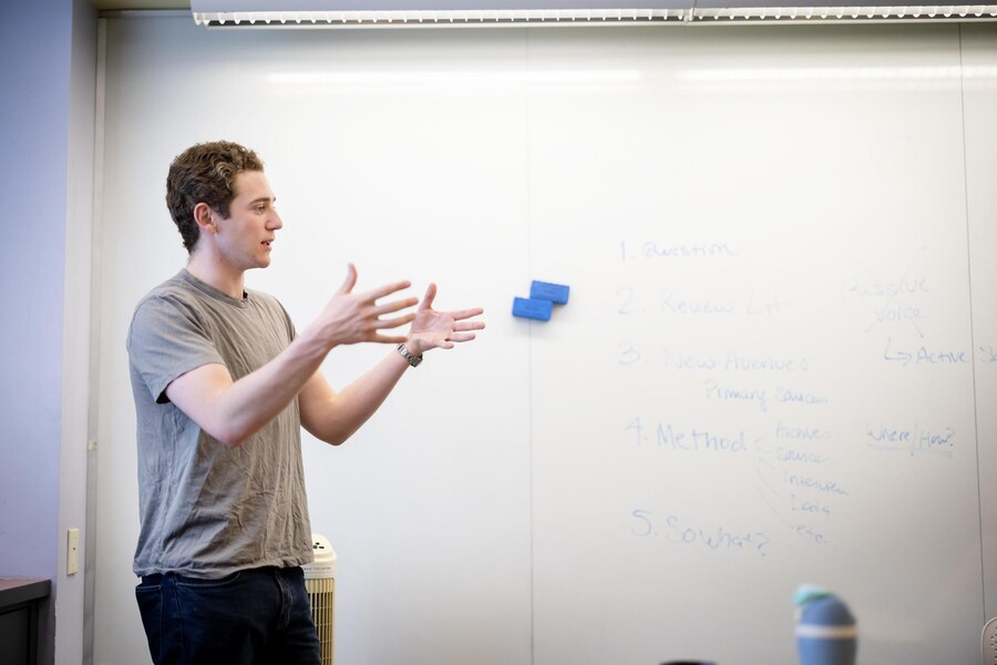 Nicholas Angellino, standing in front of a white board, gestures while presenting in class.