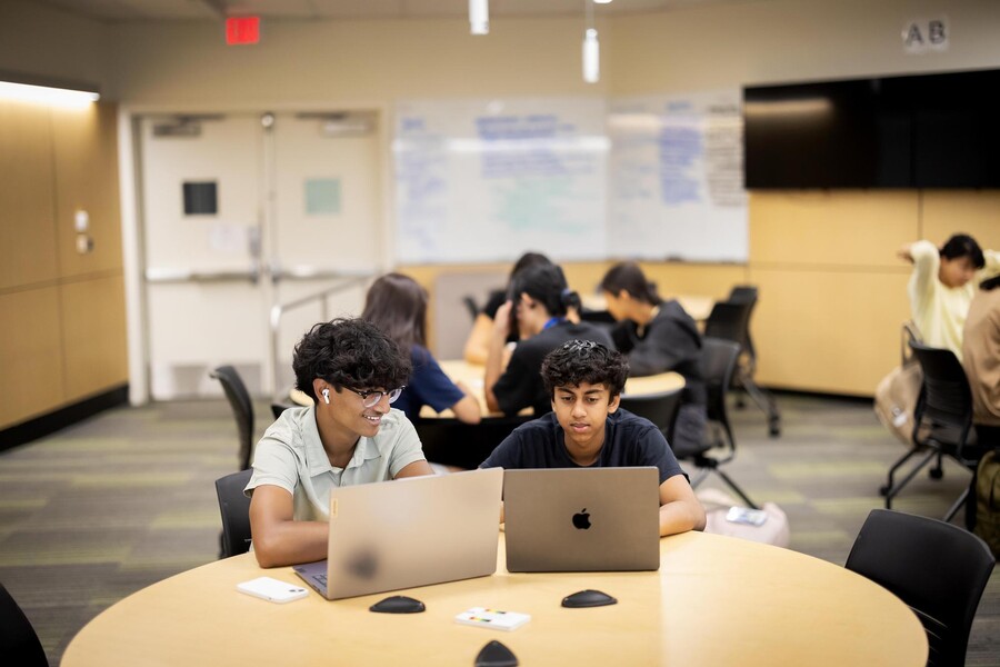 Two students sit at a table working on their laptops.