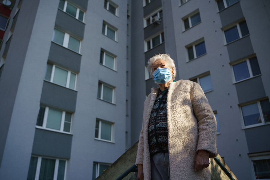 An elderly person wearing a mask outside of an urban apartment building.