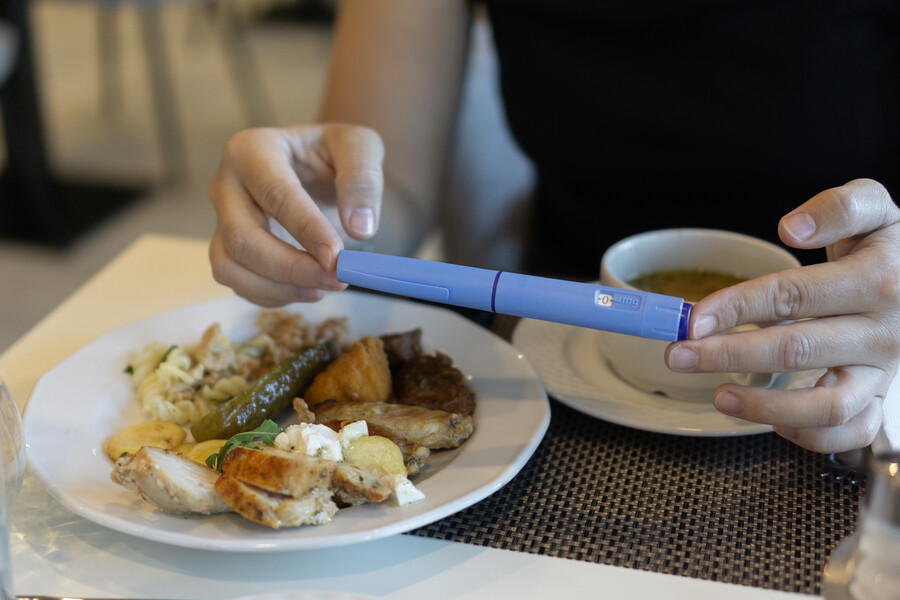 A person seated at a table holding a semaglutide pen over a plate of food.