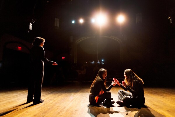 Three students practicing their One Acts scene. On the left, a student is speaking to the audience and using hand motions. On the right, two students are sitting criss-cross style, smiling and laughing while playing a patty-cake hand game.