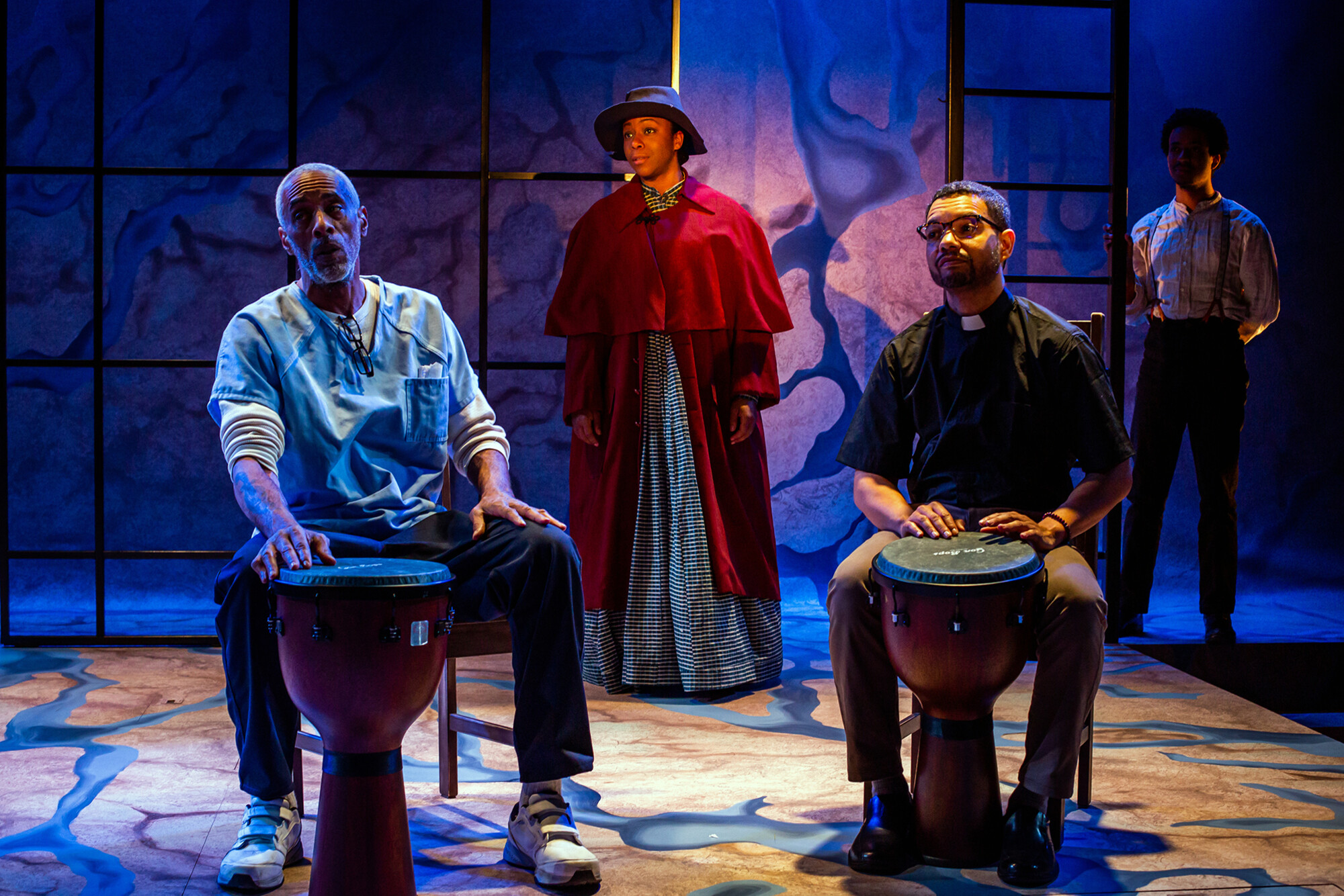 Damien j. wallace, danielle leneé, dax richardson, and aaron bell perform on stage, wallace and richardson seated in foreground playing large drums, leneé stands in background wearing a cape and hat.
