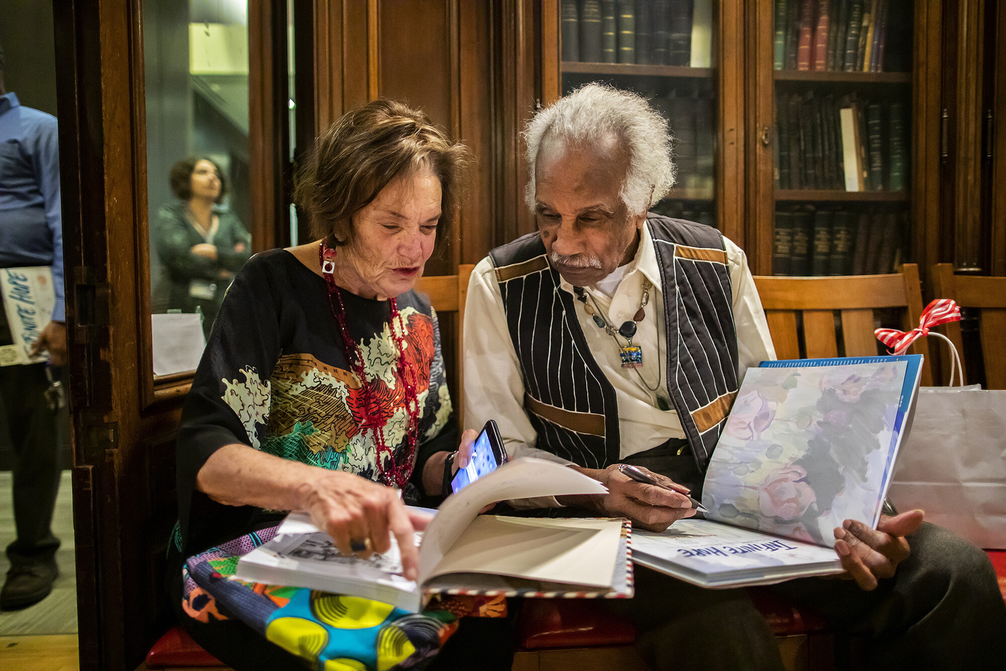 Ashley Bryan seated with an associate, both looking at two of his open books.