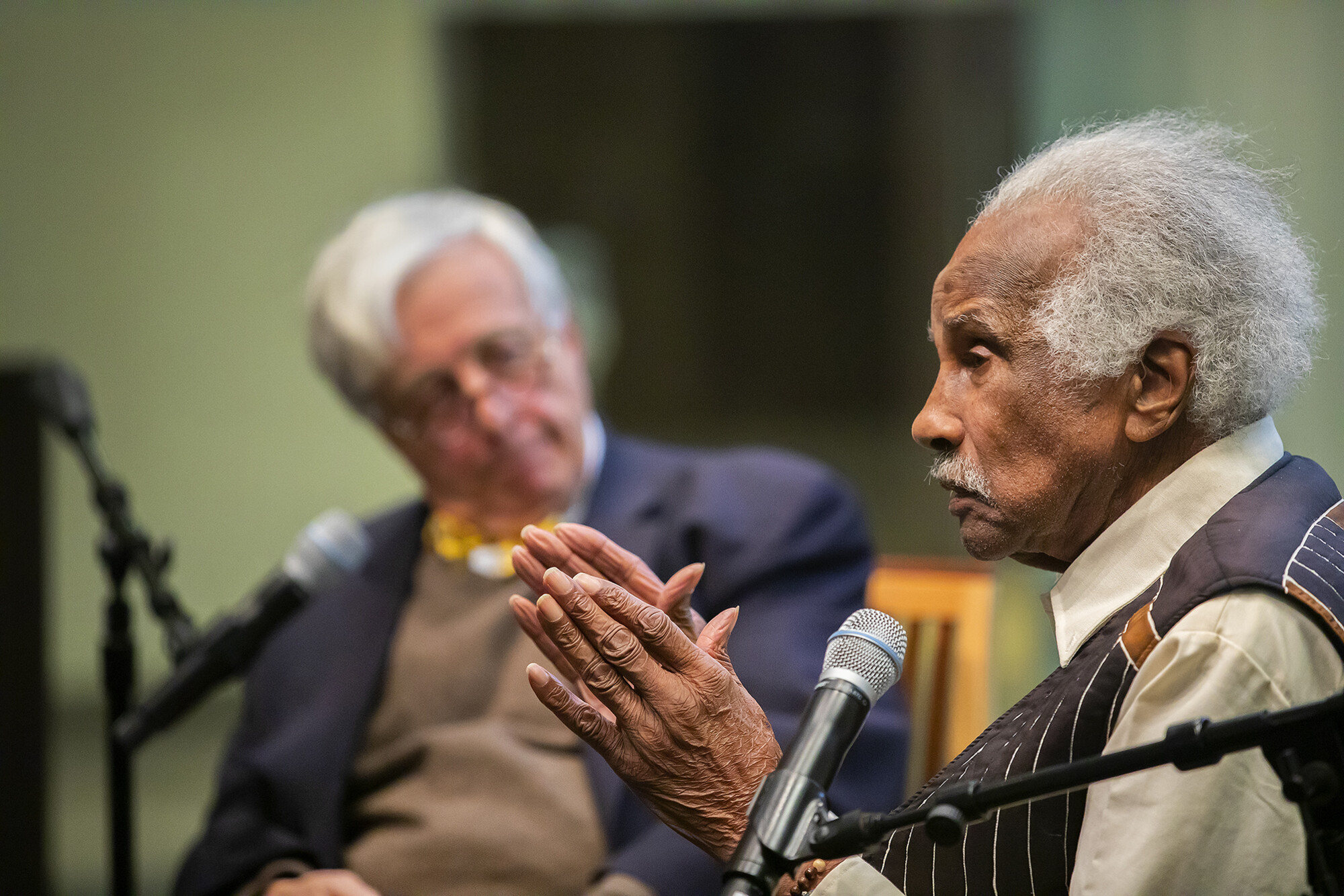 Ashley Bryan speaks into a microphone with Nick Clark looking on in background.