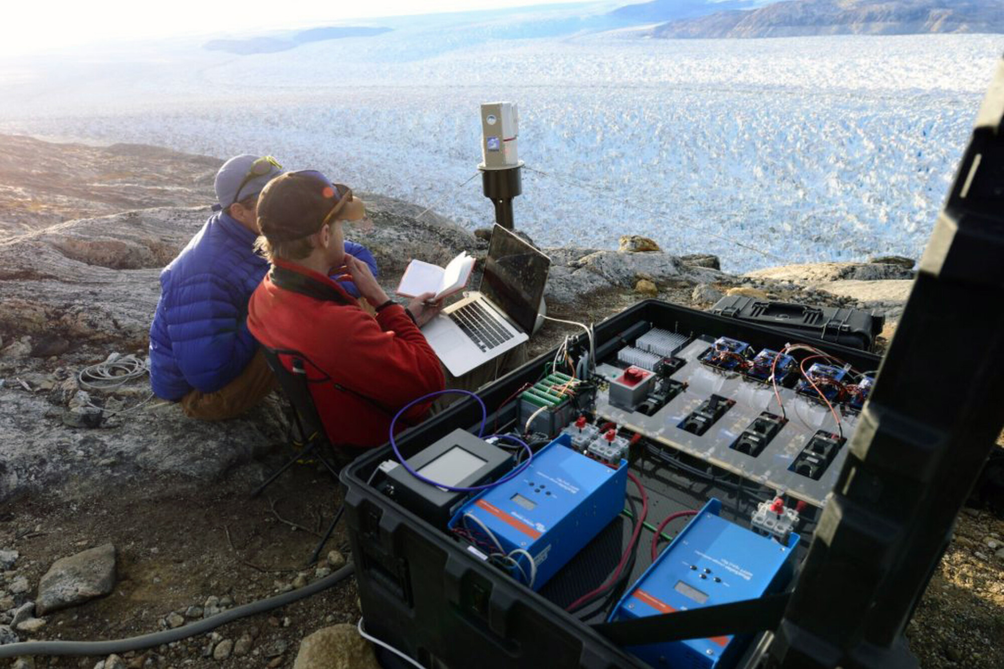 Two researchers seated with equipment overlooking a glacier.