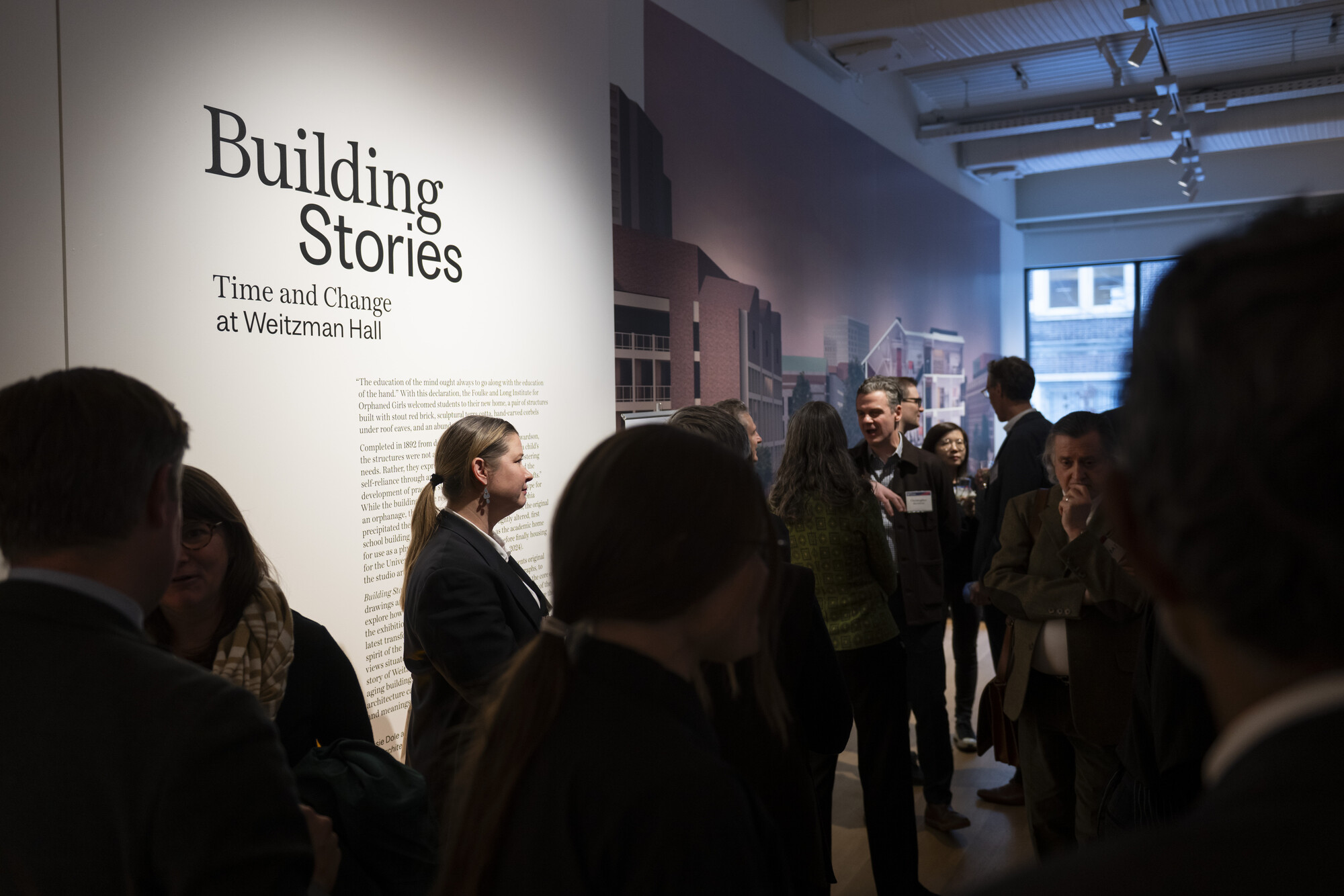 The reception for the opening of Weitzman hall, people standing by a wall with the text “Building Stories: Time and Change at Weitzman Hall.”