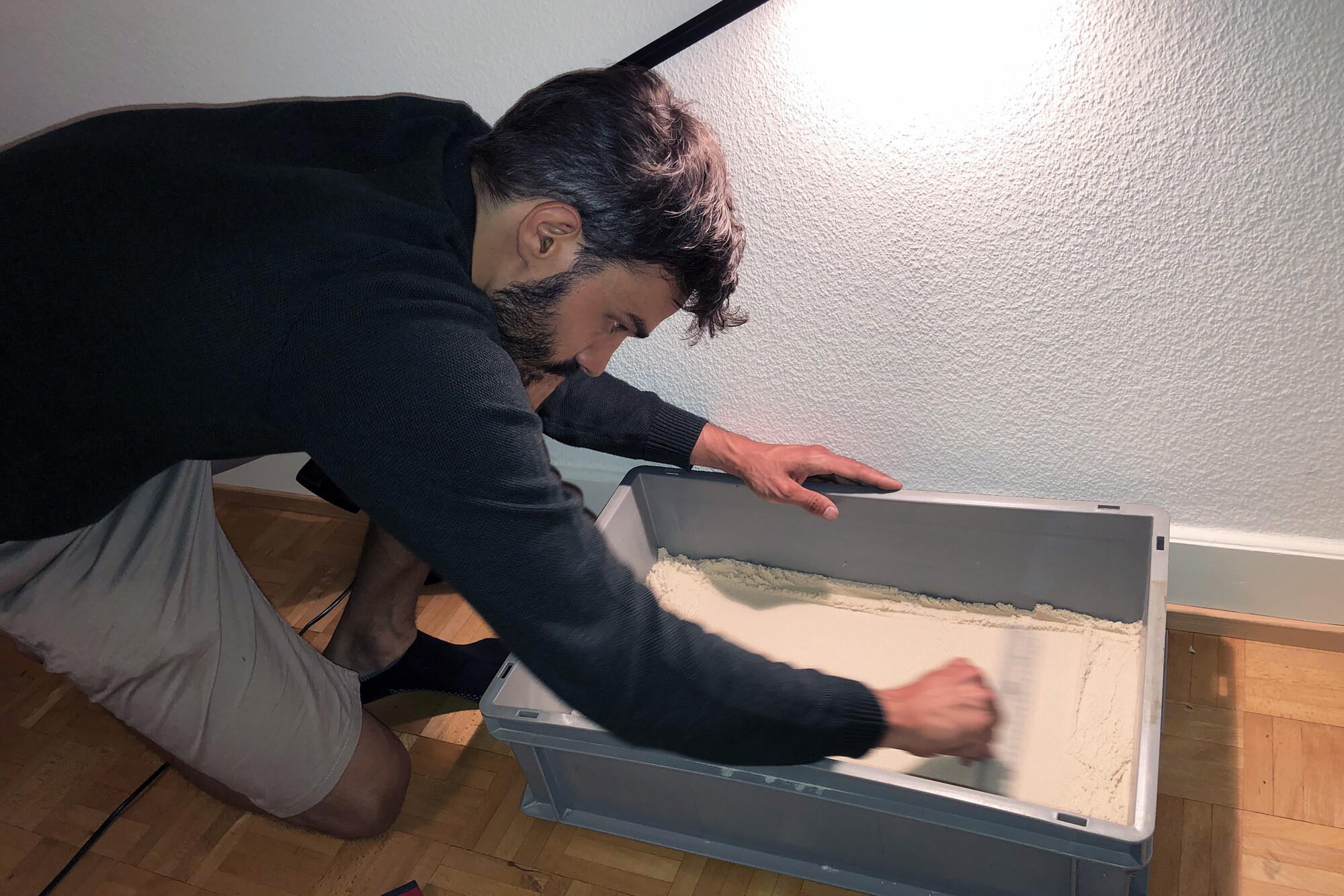 A researcher smooths a sand-filled container by hand, preparing a sloped surface for raindrop experiments.