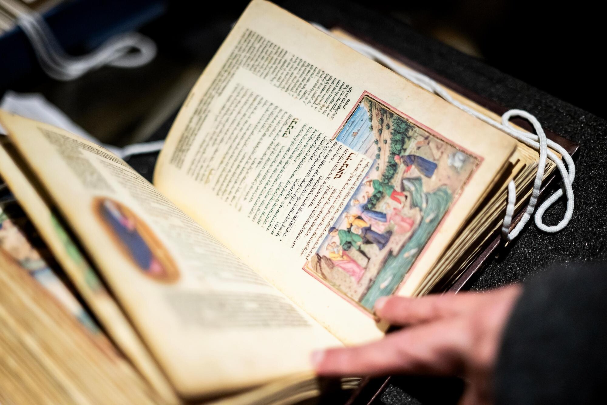 The Rothschild Miscellany, with a hand opened to a specific page with some yellowing, and featuring both Hebrew writing and vivid illustrations.