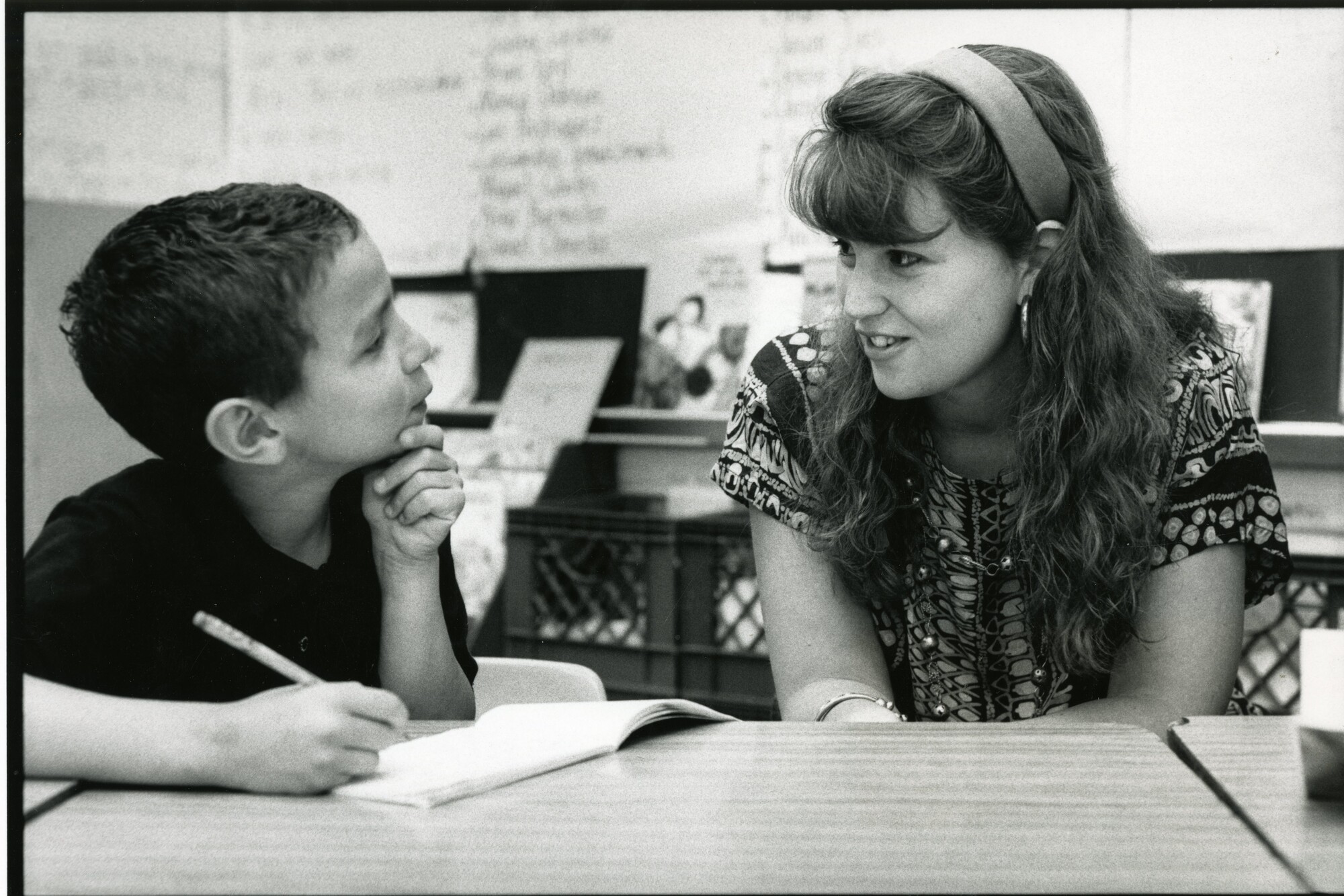 An elementary school student (left) and Penn affiliate (right) are interacting while the student writes in his booklet, in a classroom setting.