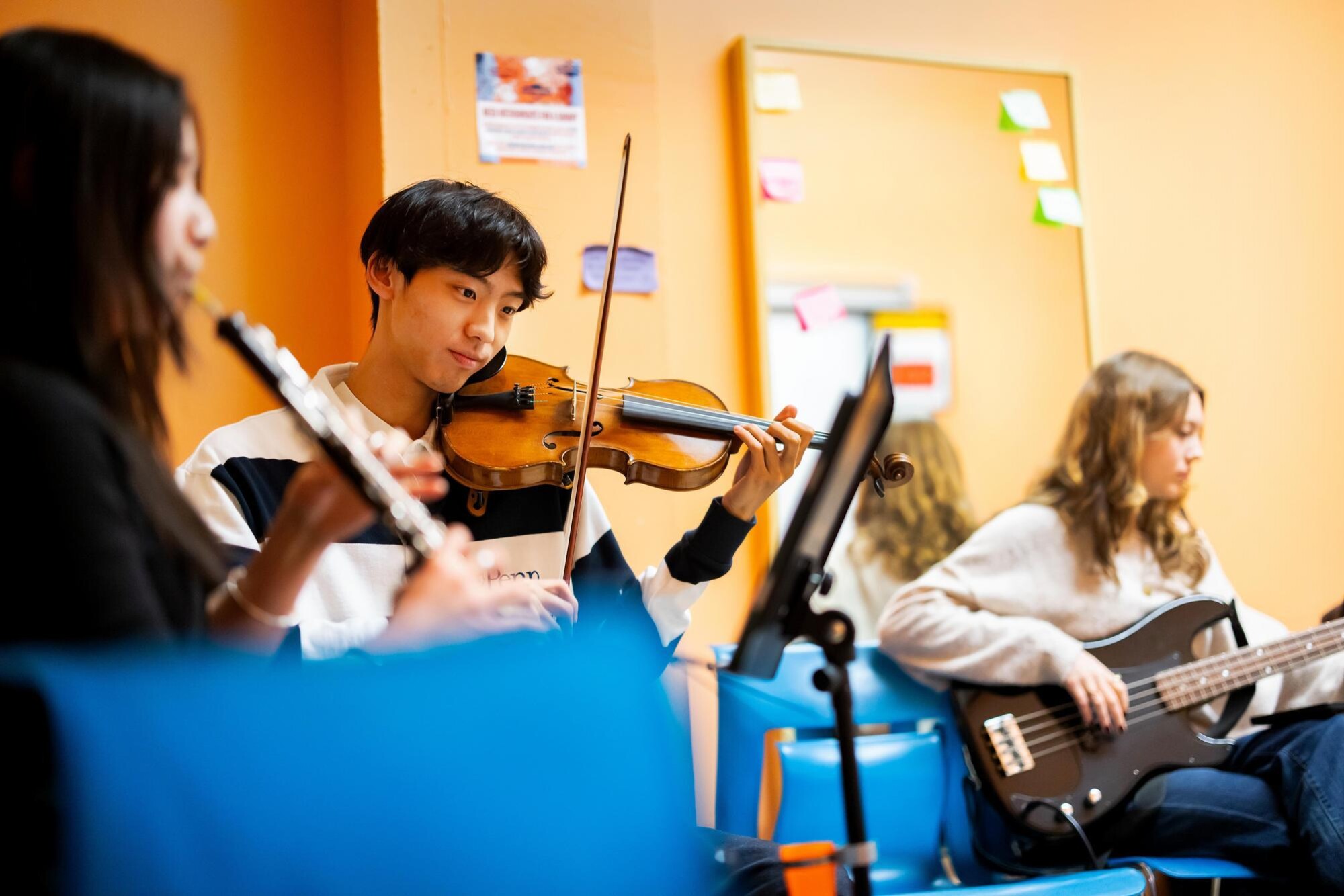 Three students in the Bloomers band group are practicing a music number. On the left, a student is playing a flute-like instrument. In the center, a student is practicing a violin-like instrument. On the right, a student is playing the guitar.