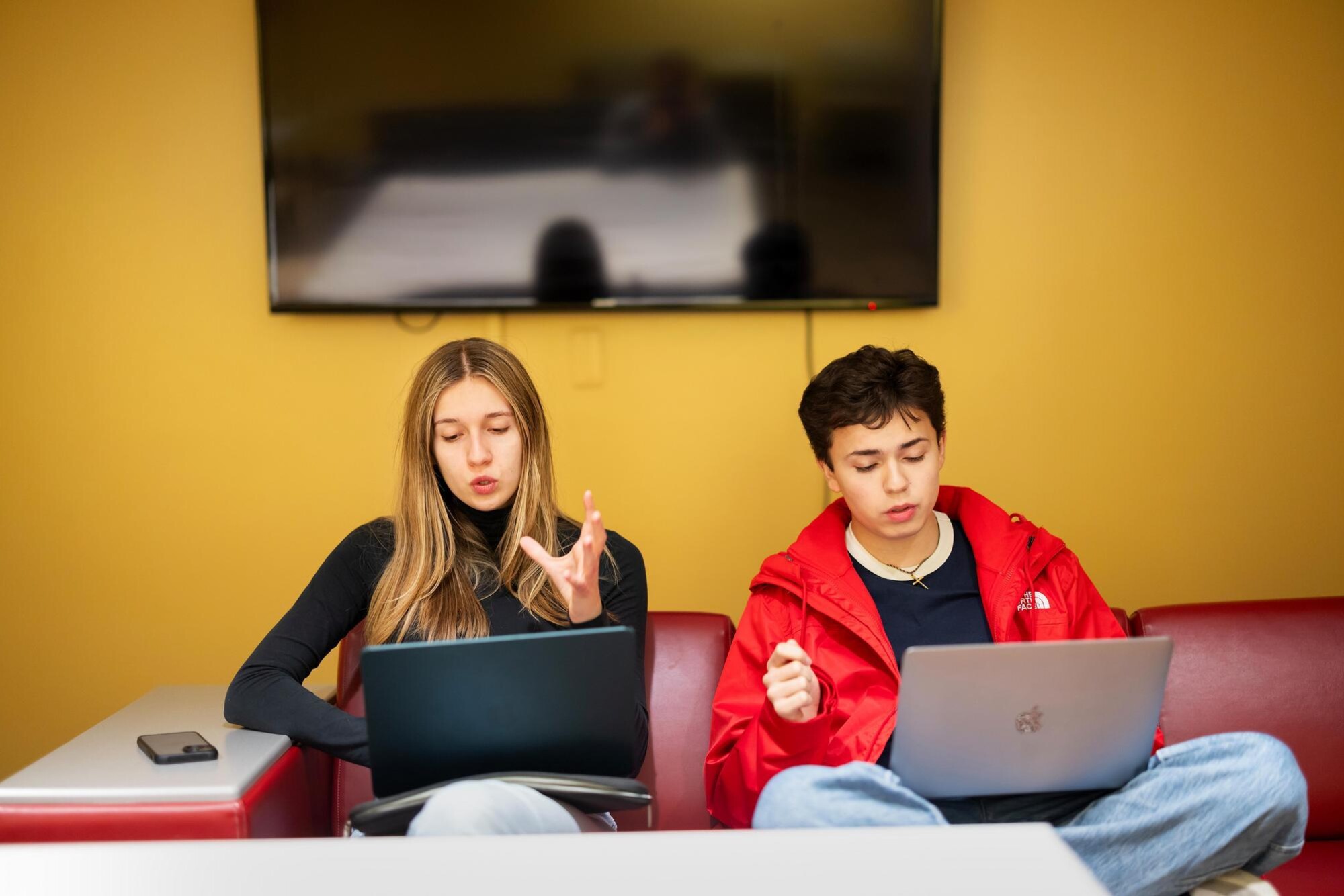 Two students in the Dischord group practicing their parts in the rehearsal number. They are sitting side-by-side on a sofa with laptops, while singing with hands raised.