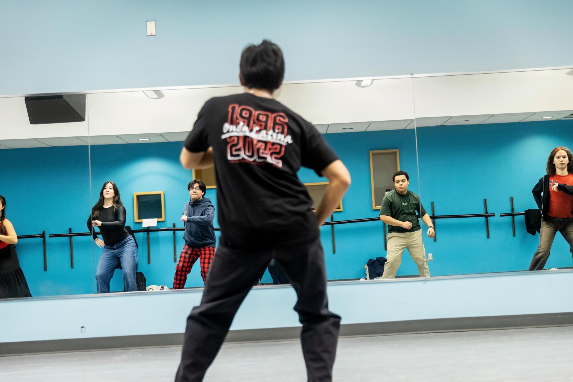 Students in the Onda Latina group practicing a dance routine. In the foreground, the group lead is dancing while guiding the group members; their shirt notes the group was established in 1996. In the background, a mirror reflects the students practicing their dance routine.