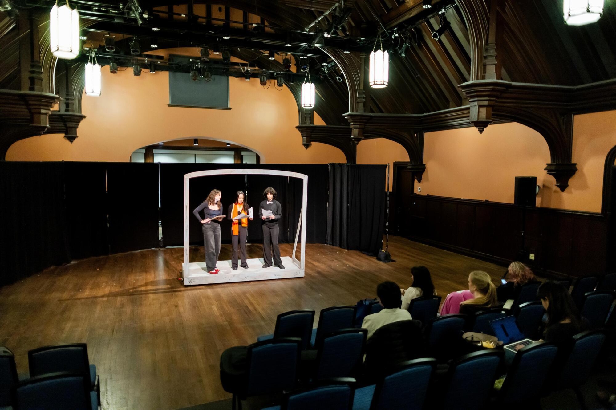 Three students practicing their One Acts scene. All three are standing under a white wooden frame and reading lines from scripts. The rehearsal audience is also pictured.