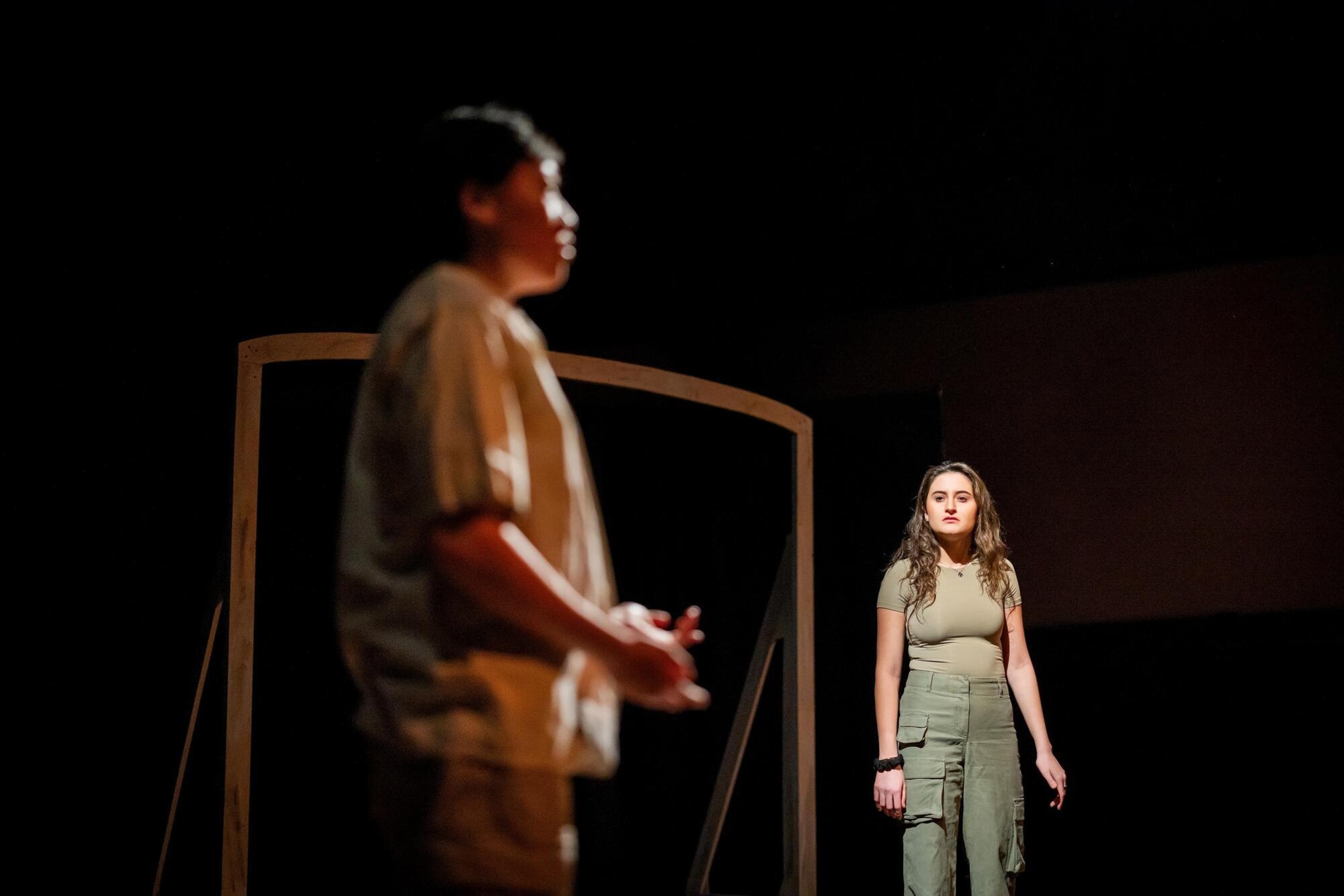 Two students practicing their One Acts scene. In the foreground, a student is speaking to the audience. In the background, a student observes the student who is speaking.