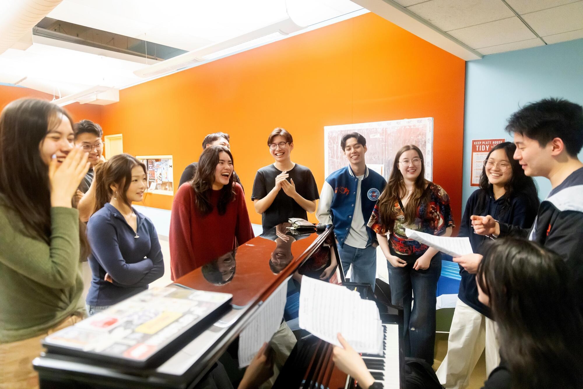 The PennYo group practicing together in unison, circled around a piano. They are laughing and smiling.