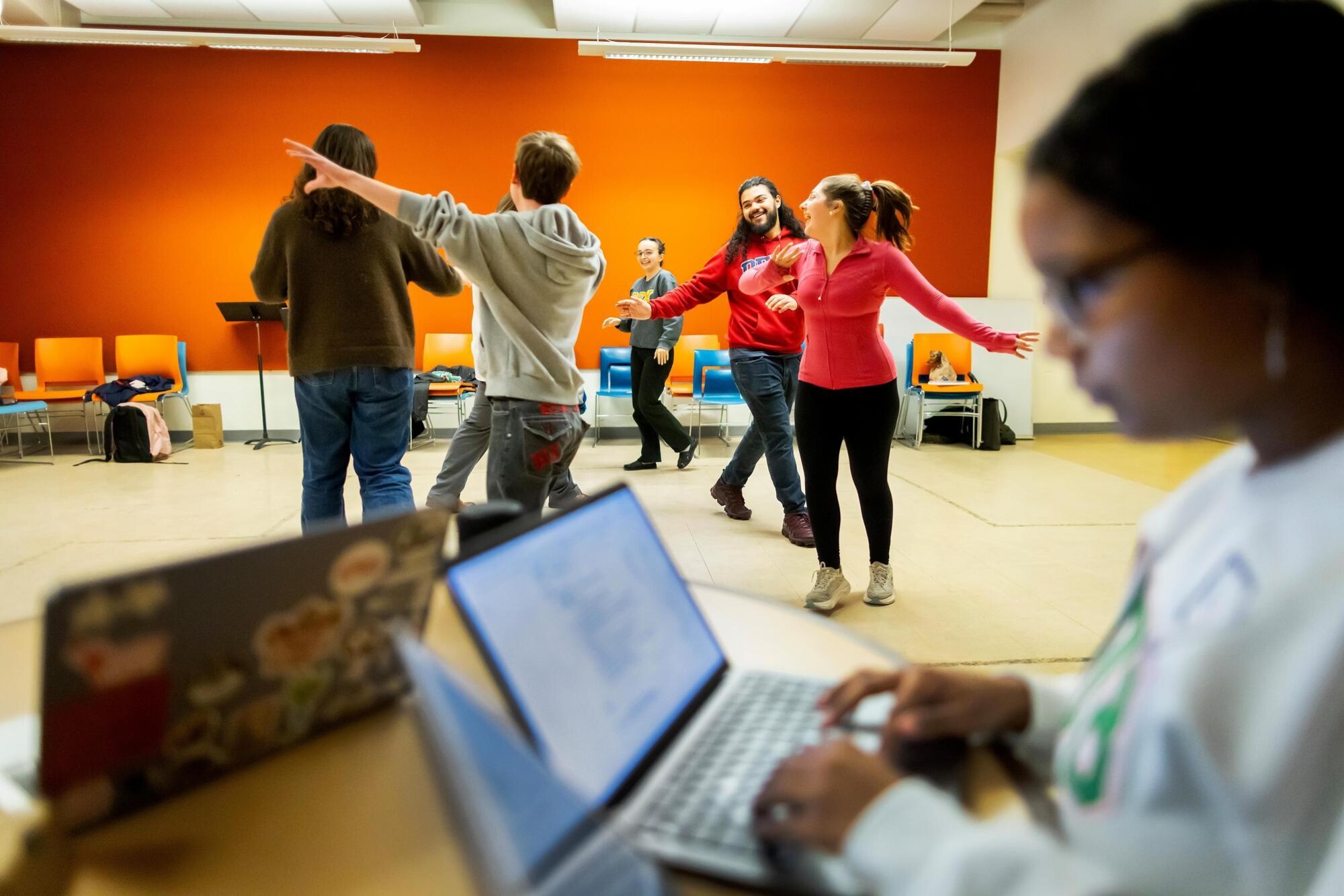 Students in the Quadramics group circling each other while rehearsing a dance routine. Their arms are extended and they are smiling and laughing. In the foreground, a student is pictured on her laptop, helping to plan the rehearsal.