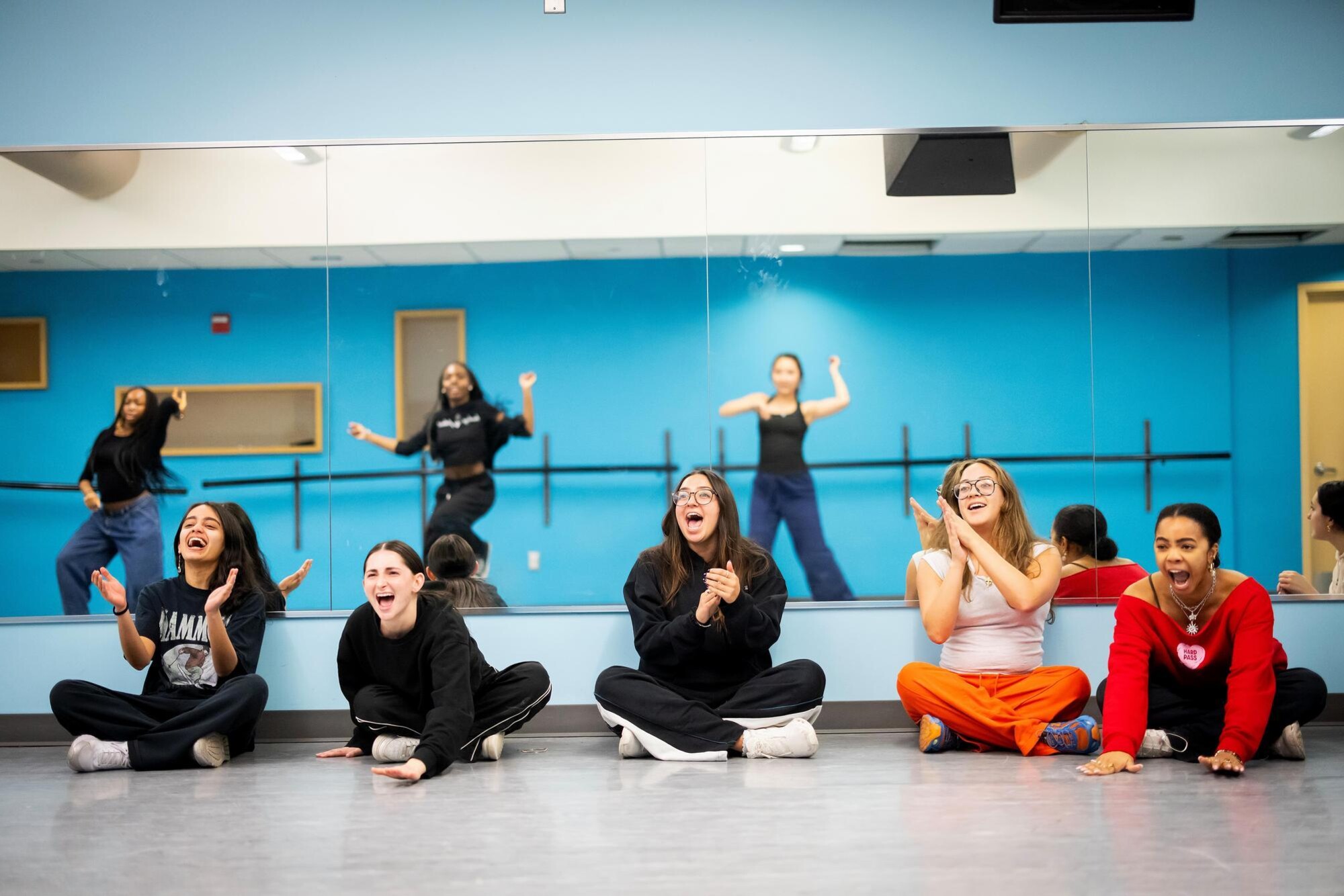 Students in the Strictly Funk group sitting next to a wall mirror. In the mirror, fellow participants are seen dancing unison. The sitting students are cheering on their peers.