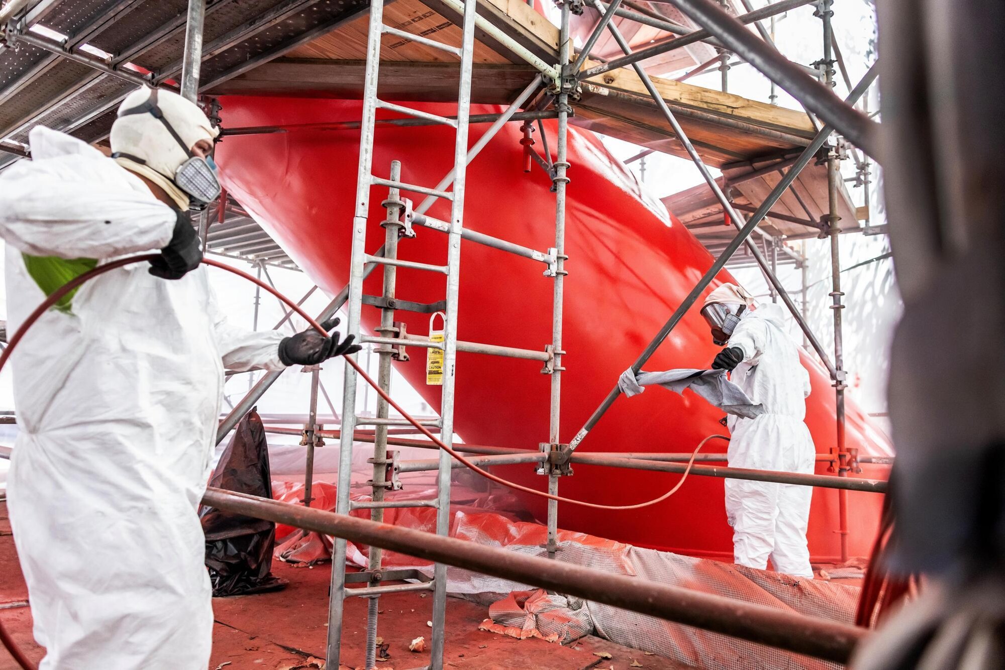 Covenant during the restoration process, newly coated in bright red and being monitored by workers in full-body protective gear. The sculpture is surrounded by scaffolding and tenting.