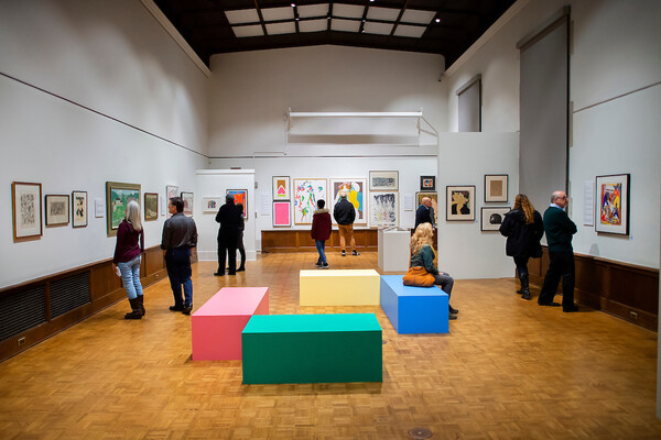 Wide view of art gallery showing many paintings on the walls and about ten people looking at them 