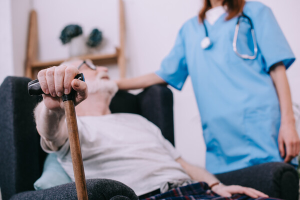 hospice nurse standing by the side of a seated elderly person holding a cane