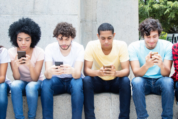 Teenagers of various races and genders sitting on a stone wall with cell phones in hand. 