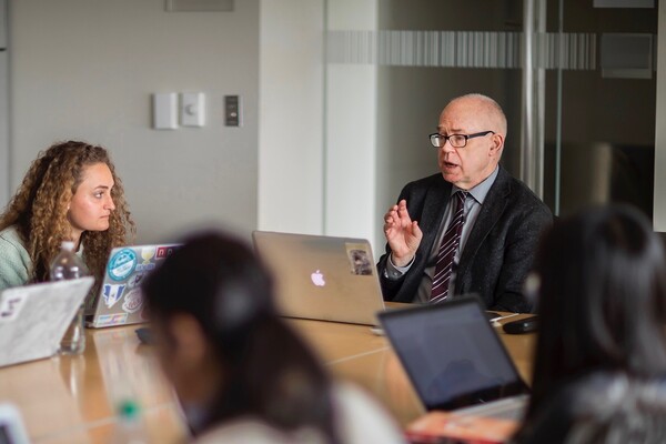 Professor speaking to classroom, sitting at table, with computer in front of him. 