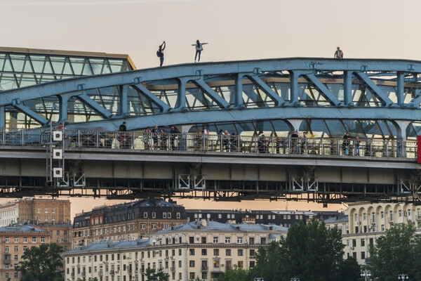 Two adolescents walking on the top structure of a bridge, a third sits, while people are on the walkway beneath the top of the bridge. 