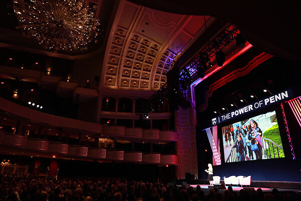 view of Amy Gutmann on stage from the audience at the Power of Penn event with a large video screen behind her showing a video