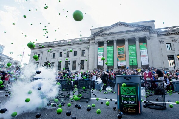 a large group of people in front of the Franklin Institute building with a science demonstration (with smoke and the aftermath of an explosion that caused colored balls to fly into the air) in the foreground