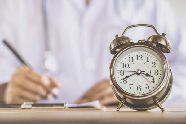 alarm clock in foreground with a doctor writing on a paper at a desk holding a pen in the background
