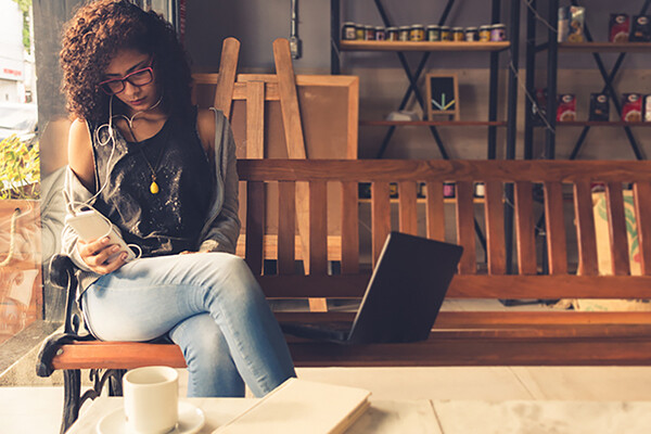 Person on a bench in a café looking at smartphone seated next to an open laptop.