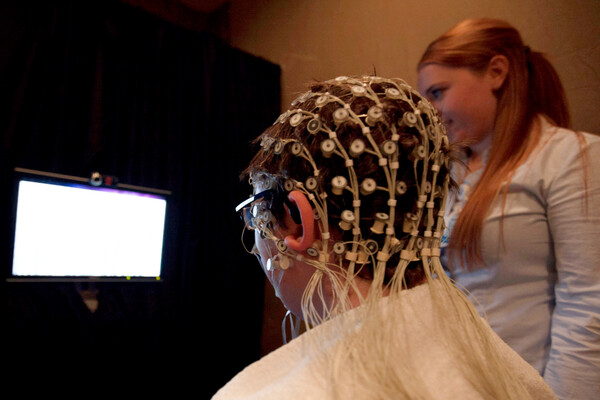 A child wearing an electroencephalogram cap looking at a bright screen, with someone standing nearby.