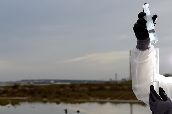 Gloved hands holding a syringe with groundwater with a background of a body of water
