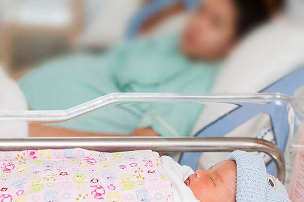 newborn baby in bassinet beside hospital bed with birth mother in background