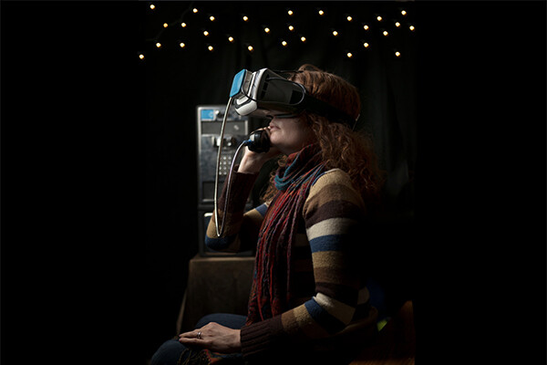 Person wearing VR glasses sitting and holding a phone to ear beside a phone booth.