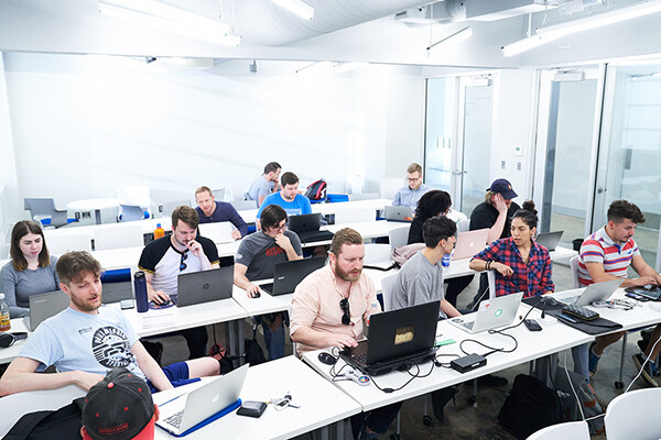 A class of adults sitting at long tables working on laptops