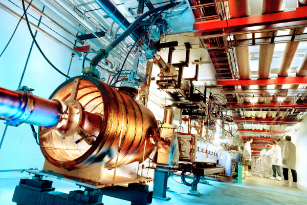a particle accelerator, a long copper tube that follows down a basement corridor below lines of tubes and wires, a group of scientists in lab coats talk with each other on the side of the room