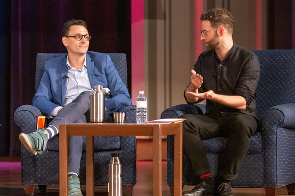 Two people sitting on a stage, one gesturing with his hands. In front of them is a brown wooden table with two water bottles.