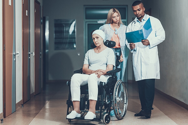 Cancer patient in wheelchair being pushed by a caregiver who consults with doctor holding a medical chart.
