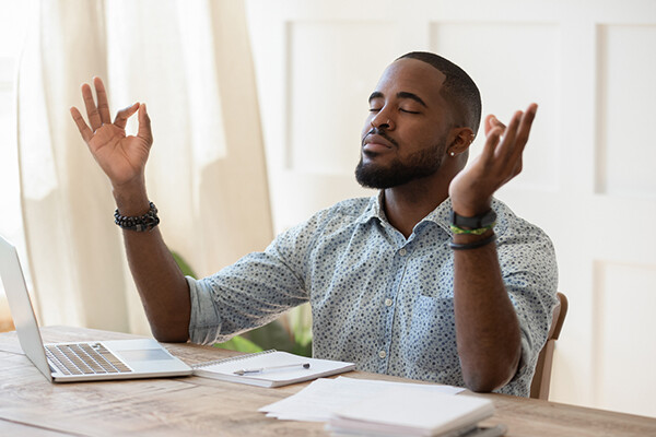 person in sunlit room with laptop and notebook with eyes closed and arms open having a meditative moment.