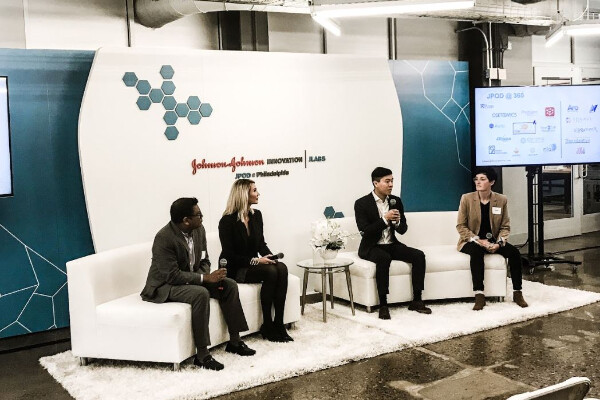 four people sitting on two couches holding microphones, one person is speaking to an audience out of view, they are next to a large screen dotted with company logos and infront of a large banner that reads Johnson and Johnson Innovation JLABS, JPOD at Philadelphia
